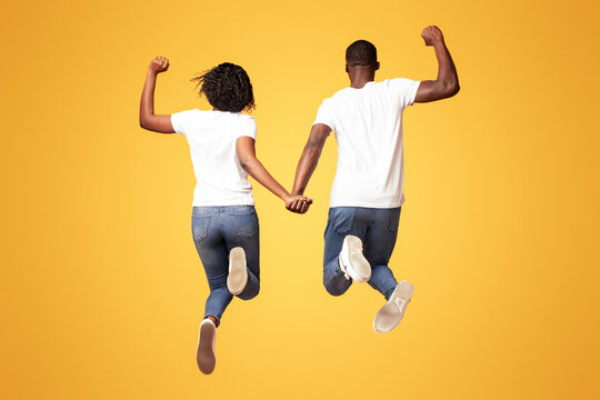Back view of african american couple in similar casual outfit holding hands and jumping up, raising fists up, celebrating success together, orange studio background, full length shot