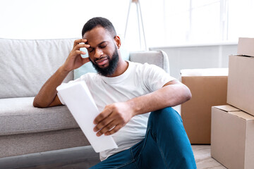 Apartment Renting Cost. Frustrated African Man Reading Paper Or Expenses Bill Sitting Among Packed...