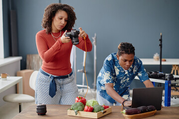 Young adult Black man using laptop while woman photographing fresh vegetables with digital camera...