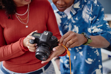 Young adult Black woman and Black man examining digital camera together, connecting orange cable to device, focusing on camera settings and technical details during food photography session