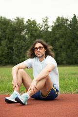 Long-haired handsome caucasian young man in sunglasses sits and relax on running track, wearing...