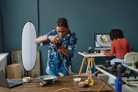 Young Black man photographing plated food on wooden table using digital camera while woman editing food images on computer in studio workspace