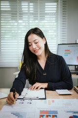 A smiling young woman reviews documents at her desk, with a laptop, calculator, and graphs on a...