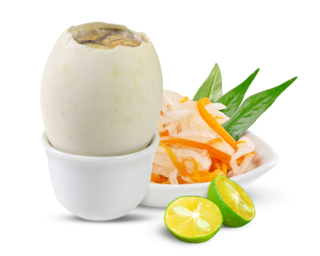 A balut egg sits in a white cup, cracked open to reveal the duck embryo, with pickled daikon, carrot, and basil beside it. Two halved calamansi limes brighten the scene against a white backdrop.