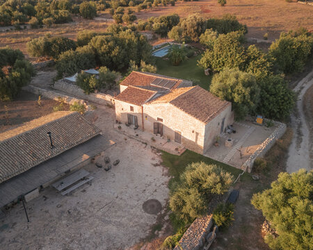 Aerial view of a rustic stone farmhouse basking in the warm glow of the setting sun, nestled among verdant trees, Rifugio Lanzagallo, Sicilia, Italy.