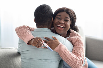 Emotional black woman wife with positive pregnancy test hugging her husband, smiling at camera,...