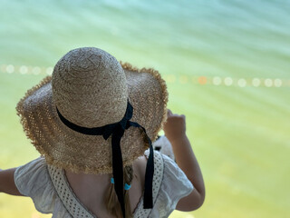 A girl in a white dress and straw hat stands by a window, looking at the calm blue sea. Childhood...