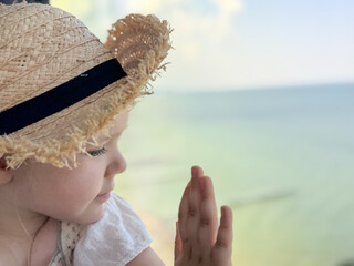 A young child in a straw hat and white dress looks out of the window at the tranquil sea, evoking...