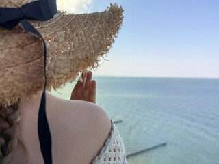 A girl in a white dress and straw hat stands by a window, looking at the calm blue sea. Childhood...