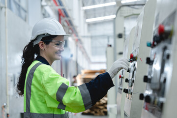 Smiling female engineer in safety gear operates industrial machine using control panel in cardboard packaging factory, showing confidence, knowledge, and safety compliance.