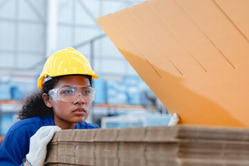 African American female worker wearing yellow helmet and safety glasses inspects stack of cardboard...