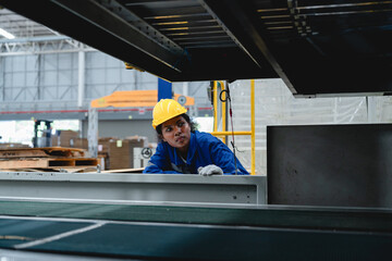 Female industrial worker wearing a yellow helmet and blue uniform examines a cardboard processing machine in a modern paper box factory, showing precision and safety in her task.