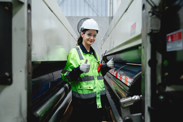 Confident female engineer wearing safety gear gives a thumbs-up gesture while standing between rollers of cardboard machinery, reflecting success and industrial readiness.
