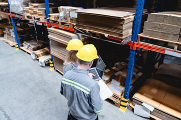 Male and female warehouse workers wearing safety helmets collaborate using laptop and clipboard while managing cardboard packaging inventory in modern factory.