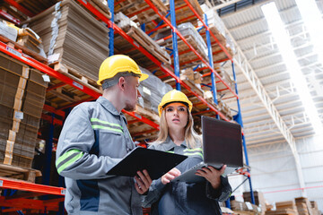 Male and female warehouse workers wearing safety helmets collaborate using laptop and clipboard while managing cardboard packaging inventory in modern factory.