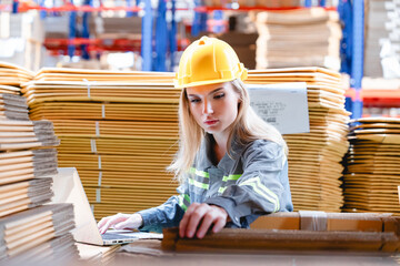 Confident female warehouse worker in safety uniform using laptop for inventory check in cardboard packaging area. Thumbs-up gesture shows successful operation in industrial storage zone.