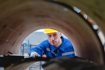 Caucasian male engineer in reflective jacket and white helmet examines mechanical components of large industrial paper machine inside cardboard packaging plant.