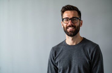 Obraz premium A smiling man with glasses and a beard wearing a casual gray sweater stands against a plain gray background