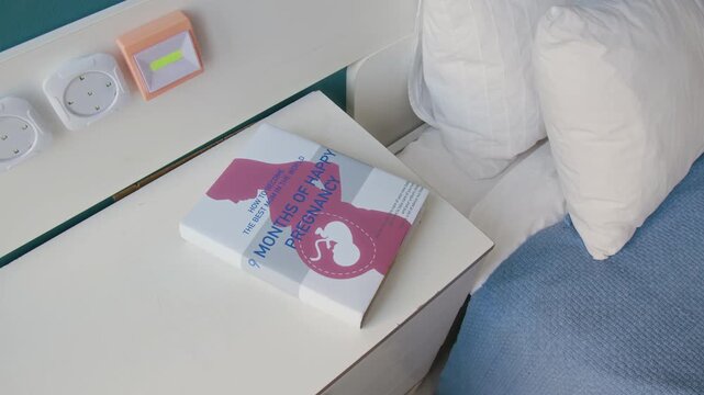 No people close-up of pregnancy reference book, cover displaying expecting mother with fetus, lying on top of dresser near bed with blue sheets and soft pillows, in natural daylight