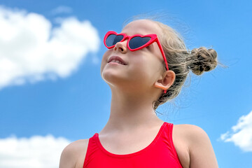 Cute little girl wearing red shirt top and heart-shaped sunglasses against a background of blue sky with white clouds. Carefree childhood, peace, love and freedom concept. High quality photo