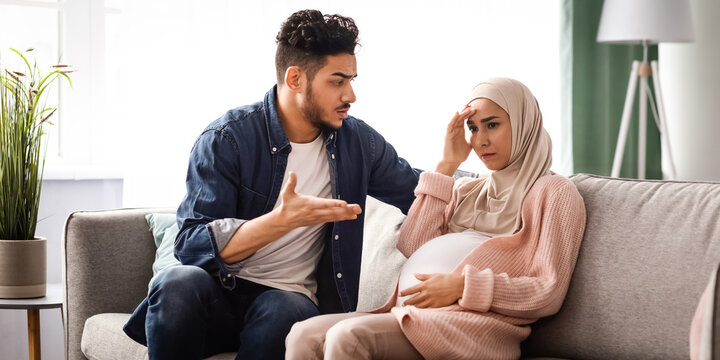 Aggressive arab man arguing with his upset pregnant muslim wife at home, sitting on couch in living room. Stressed expectant lady in hijab feeling unwell, tired of domestic conflicts and quarrels