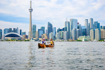 Fototapeta premium Active outdoor lifestyle: young adult couple paddling a canoe on the Toronto Islands in summer with the downtown city skyline in the background room for text copy space