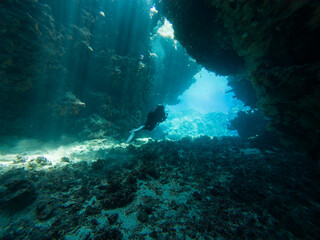 Landscape view of sunlight sunbeams dancing from the surface in underwater sea cave with rock walls and scuba diver exploring