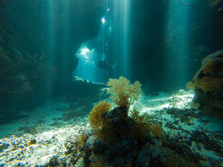 Landscape view of sunlight sunbeams dancing from the surface in underwater sea cave with rock walls and scuba diver exploring