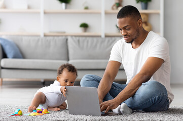 Freelancing With Baby. Young African American Man Working On Laptop And Taking Care Of His Infant Child At Home, Black Father Using Computer For Online Work And Babysitting Toddler Kid, Copy Space