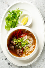 Bowl of Vietnamese pho soup with beef, rice noodles, green onions, cilantro, and lime wedges on a textured surface, showcasing traditional Asian cuisine.