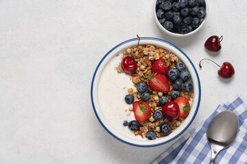 Natural yogurt with granola, blueberries and strawberries in bowl on light background with spoon and napkin. Healthy and nutritious breakfast concept. Top view and copy space.