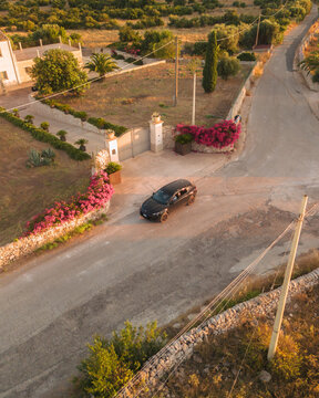 Aerial view of a sleek black car pauses before an ornate gate adorned with vibrant bougainvillea, amidst sun-kissed fields, Ispica, Sicily, Italy.