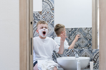 Young boy with shaving cream on his face sits at a sink in a contemporary bathroom, expressing surprise and fun while playing. Light colors enhance the playful atmosphere.