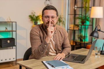 Shh be quiet please. Caucasian young businessman presses index finger to lips makes silence gesture sign do not tells secret at home office desk. Adult man with laptop in living room in apartment.