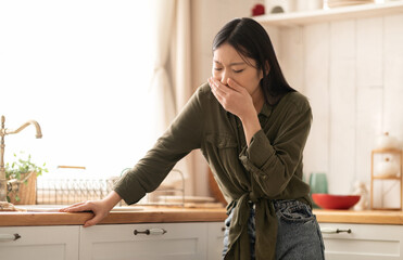 Young asian woman in casual feeling nausea, cover mouth, standing next to table in kitchen, copy...