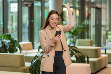 Happy amazed young businesswoman girl working on smartphone shocked by sudden victory game winning...
