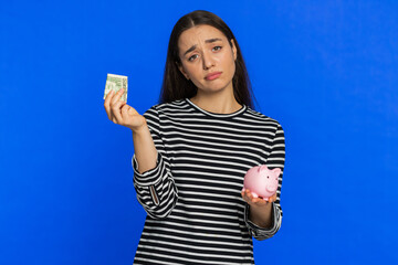 Poor Caucasian woman in striped blouse insufficient amount of money, holding piggybank and one...