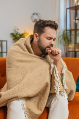 Sick Caucasian young man covered with blanket coughing while sitting on couch in living room at...