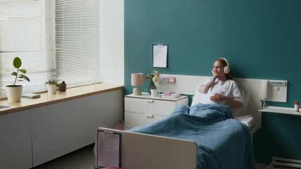 Wide shot of happy, smiling pregnant young Caucasian woman in white medical gown relaxing in bed at maternity ward, listening to music with headphones and caressing baby in stomach - Powered by Adobe