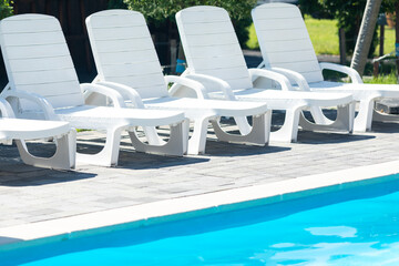 Empty poolside with white sun loungers and blue water. Row of white plastic sunbeds next to swimming pool on hotel outdoor patio. Summer vacation concept