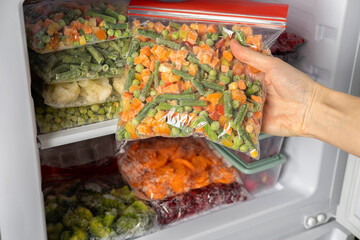 Female hands retrieving frozen vegetable mix from freezer. Plastic bags with different frozen vegetables and berries in refrigerator.  Winter supply food in zip bags, preserving nutrients.