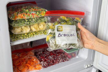 Female hands retrieving frozen broccoli from freezer. Plastic bags with different frozen vegetables and berries in refrigerator.  Winter supply food in zip bags, preserving nutrients.