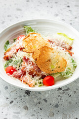 Fresh Caesar salad with crispy bacon, cherry tomatoes, Parmesan cheese, and toasted bread slices served in a white bowl on a speckled gray surface.