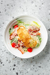 Fresh Caesar salad with crispy bacon, cherry tomatoes, Parmesan cheese, and toasted bread slices served in a white bowl on a speckled gray surface.