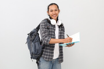 Young Asian male student wearing casual clothes and backpack, headphones around neck, smiling looking at camera while writing in book, standing isolated on white background.