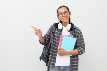 Young asian student wearing casual clothes, backpack, glasses, headphones around neck, carrying books, pointing sideways at empty space, smiling looking at camera, isolated on white background.