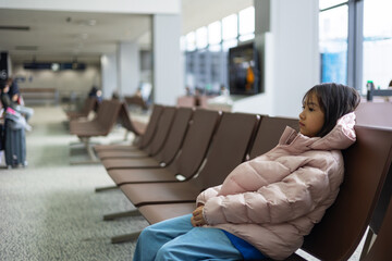 Asian adorable kid girl traveler in winter jacket sits alone in airport lounge, lost in thought. The stillness, vacant chairs, and soft lighting capture solitude, patience, and the journey ahead.