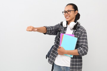 Handsome asian student man wearing casual clothes, backpack, glasses, headphones around neck, gesturing thumbs up, smiling facing side in empty space, standing isolated white background.
