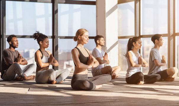 Meditation Session. Group Of Diverse Sporty People Practicing Yoga In Modern Gym, Sitting In Lotus Position With Namaste Gesture, Side View - Powered by Adobe