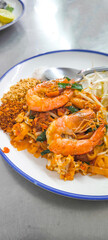 Pad Thai with fresh shrimp in a white plate with blue edges on a stainless steel table.